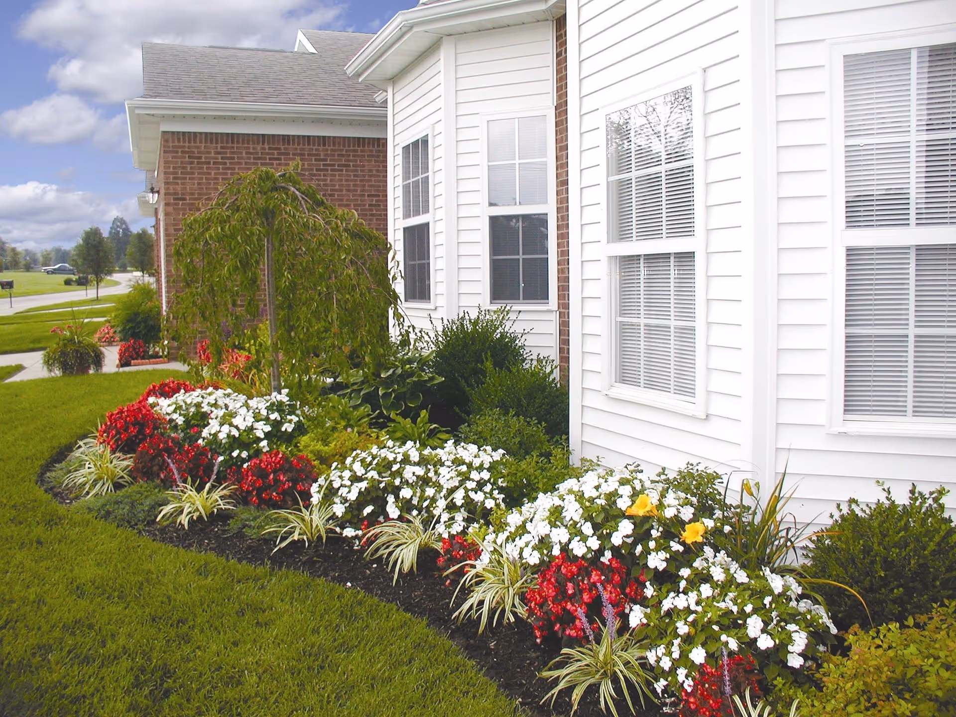 A well-maintained garden bed with red, white, and yellow flowers and green shrubs along the side of a building with white siding and brick exterior. The lawn is neatly mowed, and there is a sidewalk and a road visible in the background under a partly cloudy sky.