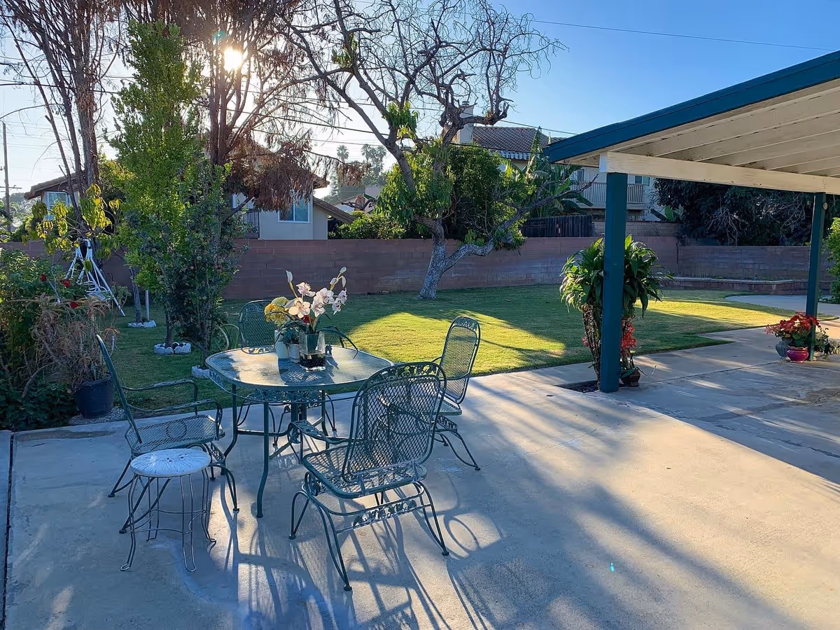 Outdoor patio area with a metal table and four chairs, decorated with a flower arrangement on the table. The patio is adjacent to a grassy lawn with trees and plants, and there is a covered structure with green posts and a white roof providing shade. The sun is shining through the trees in the background.