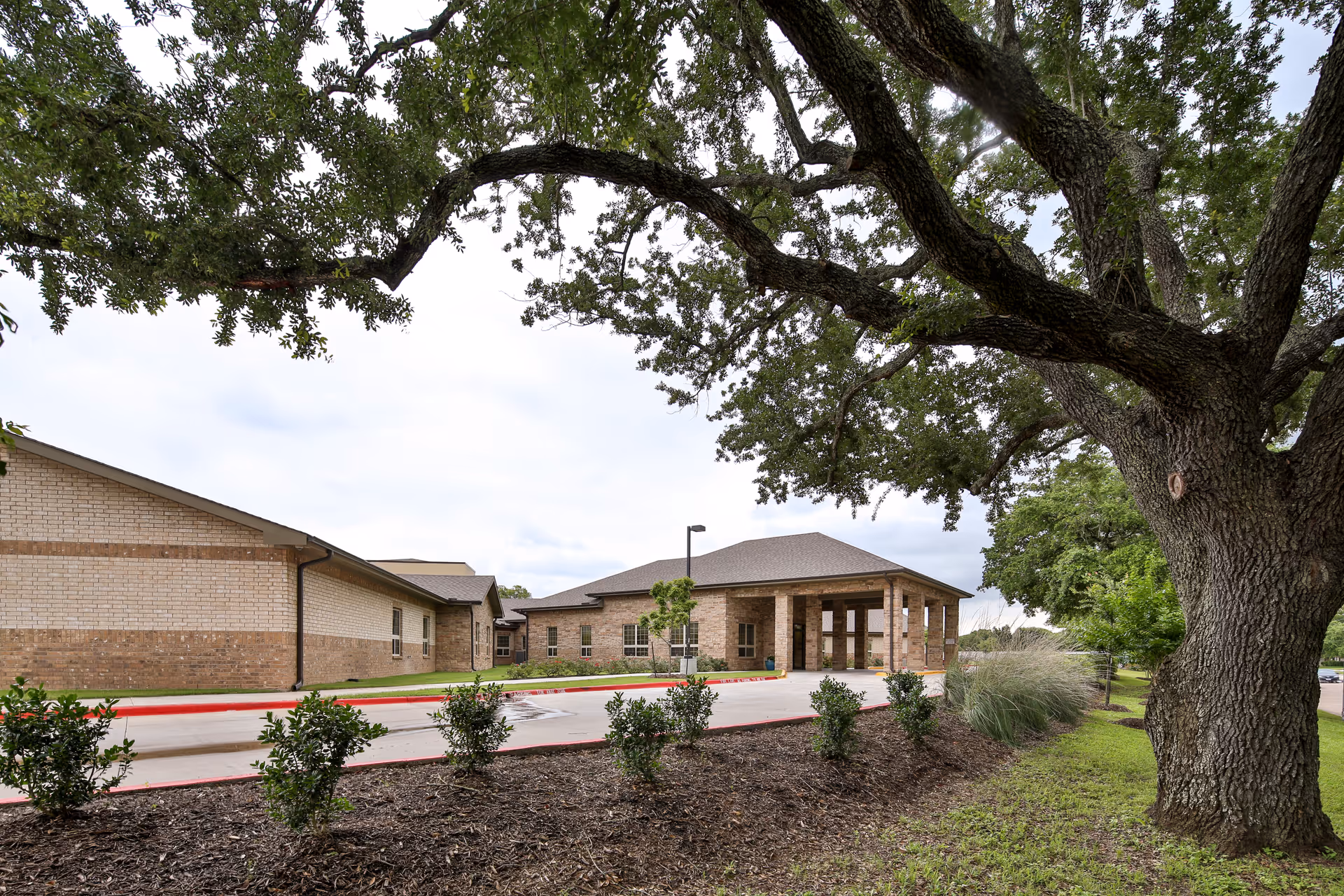 Exterior view of a single-story brick building with a covered entrance, surrounded by landscaping including bushes, grass, and a large tree in the foreground.