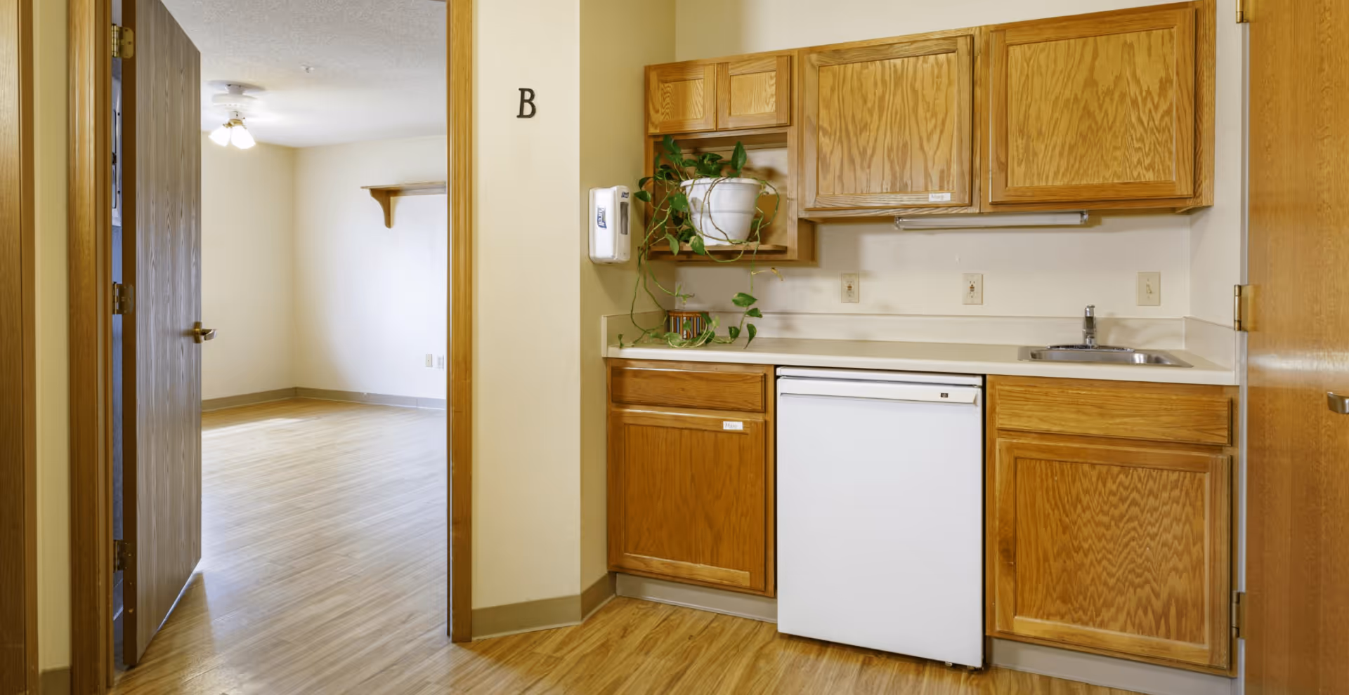 Interior view of a senior living facility unit showing a small kitchenette with wooden cabinets, a white mini refrigerator, a sink, and a potted plant on a shelf. To the left, there is an open doorway leading to an empty room with wooden flooring and a ceiling light fixture.