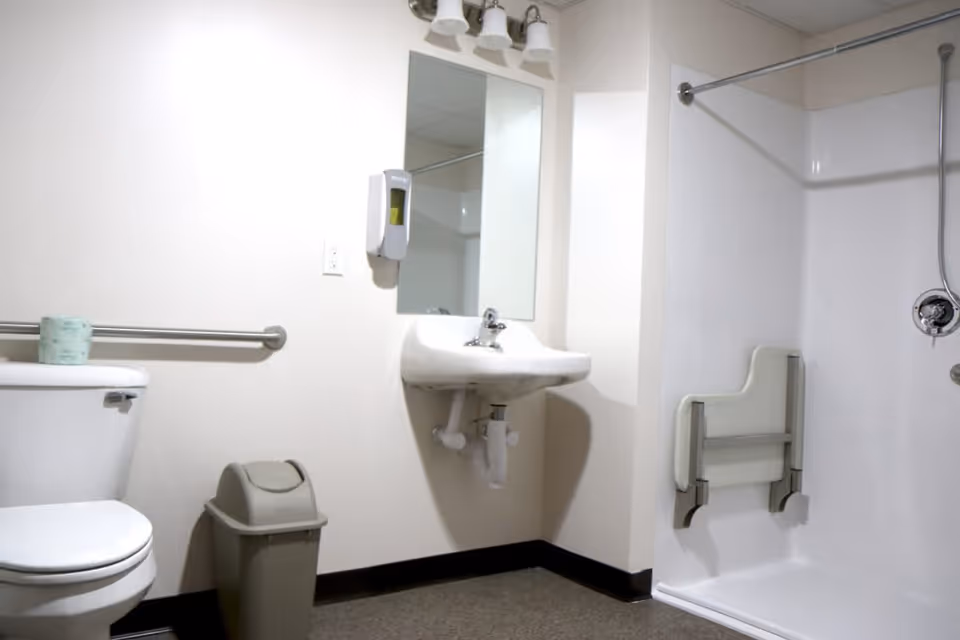 A clean, accessible bathroom with a white toilet, a small trash bin, a wall-mounted sink with a mirror above it, a soap dispenser on the wall, and a shower area with a foldable seat and grab bars.