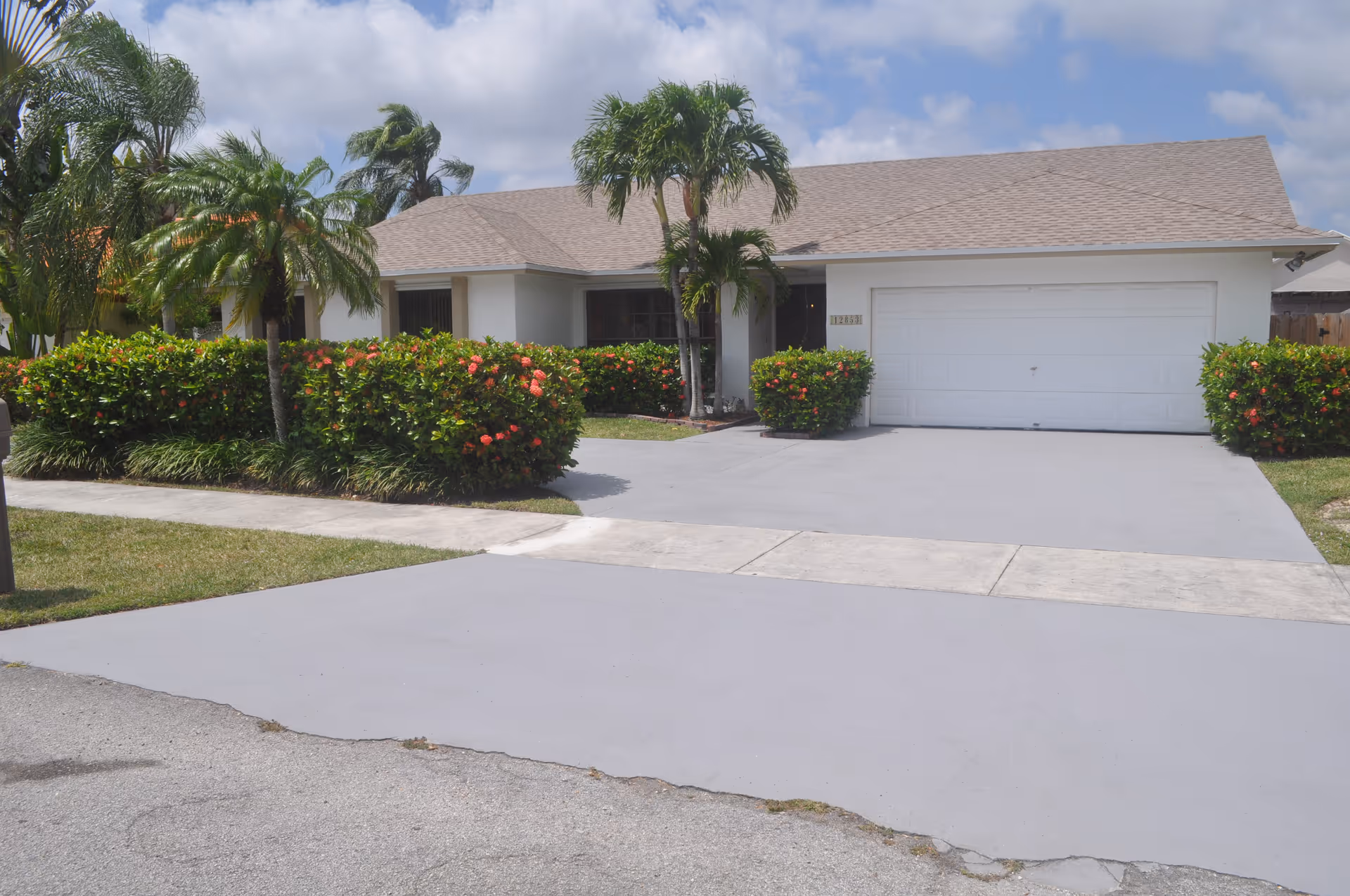 Single-story house with a beige roof, white exterior walls, and a two-car garage. The driveway and sidewalk are visible in front of the house. There are palm trees and green bushes with red flowers in the front yard under a partly cloudy sky.