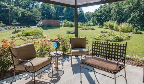 Outdoor seating area under a covered patio with cushioned chairs and a bench, overlooking a landscaped garden with green grass, flower beds, and trees in the background.