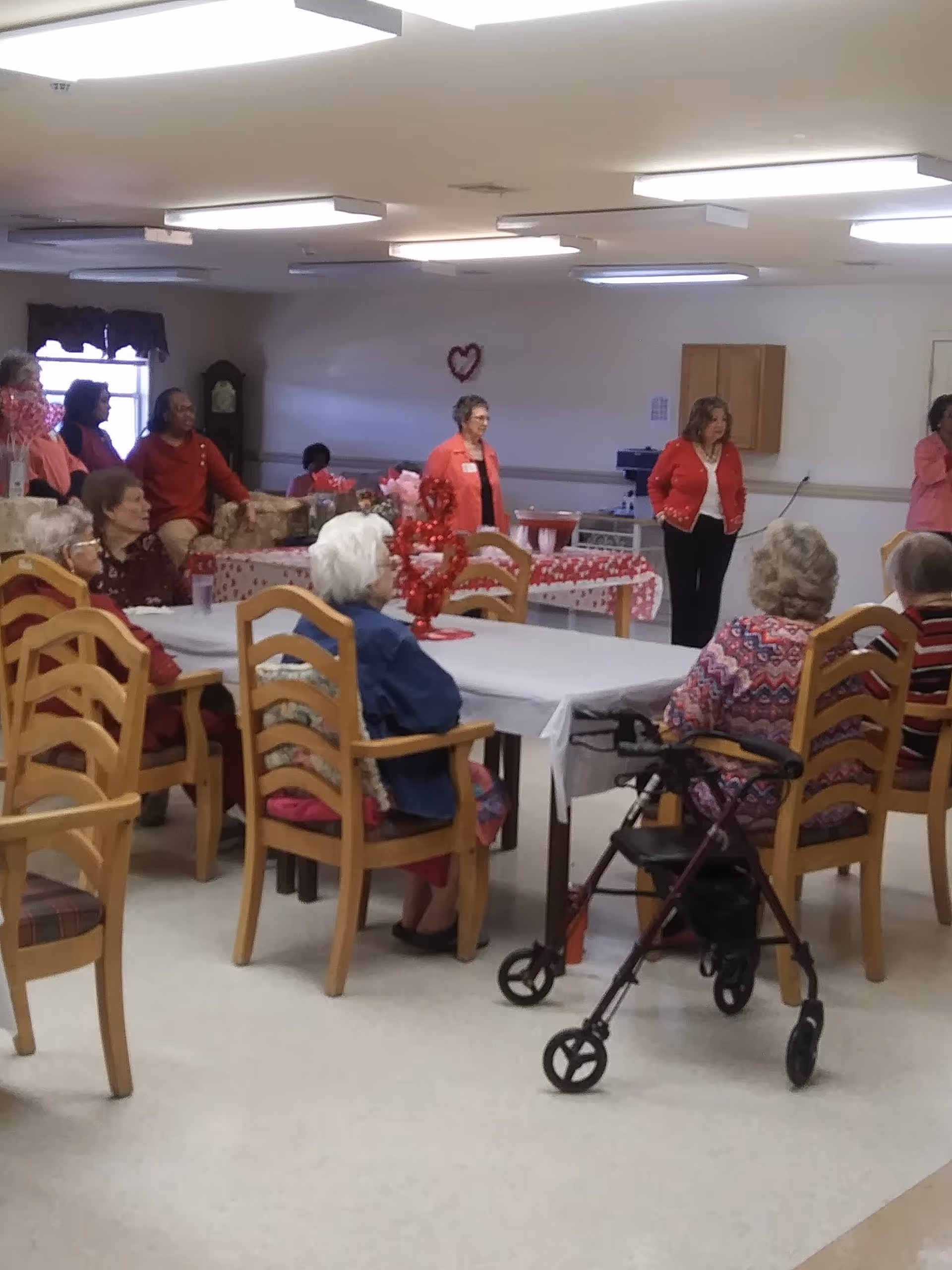 A group of elderly people seated around tables in a community room decorated with red and pink Valentine-themed decorations. Several women are standing near the back wall, some wearing red clothing. A walker is positioned near one of the seated individuals.