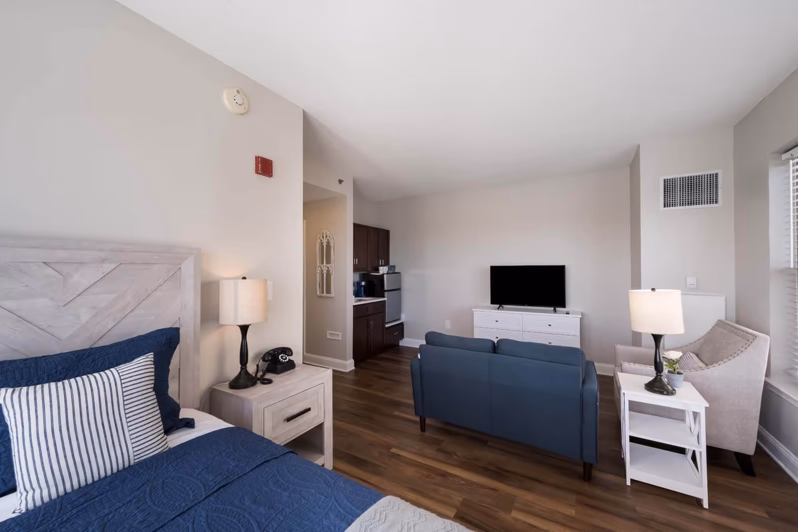 A bright and modern studio apartment featuring a bed with blue bedding and striped pillows on the left, a nightstand with a lamp and a vintage telephone, a small sitting area with a blue loveseat and a beige armchair, a white side table with a lamp and a small plant, a flat-screen TV on a white dresser, and a kitchenette with dark cabinets and a mini fridge in the background. The room has wood flooring and light-colored walls.