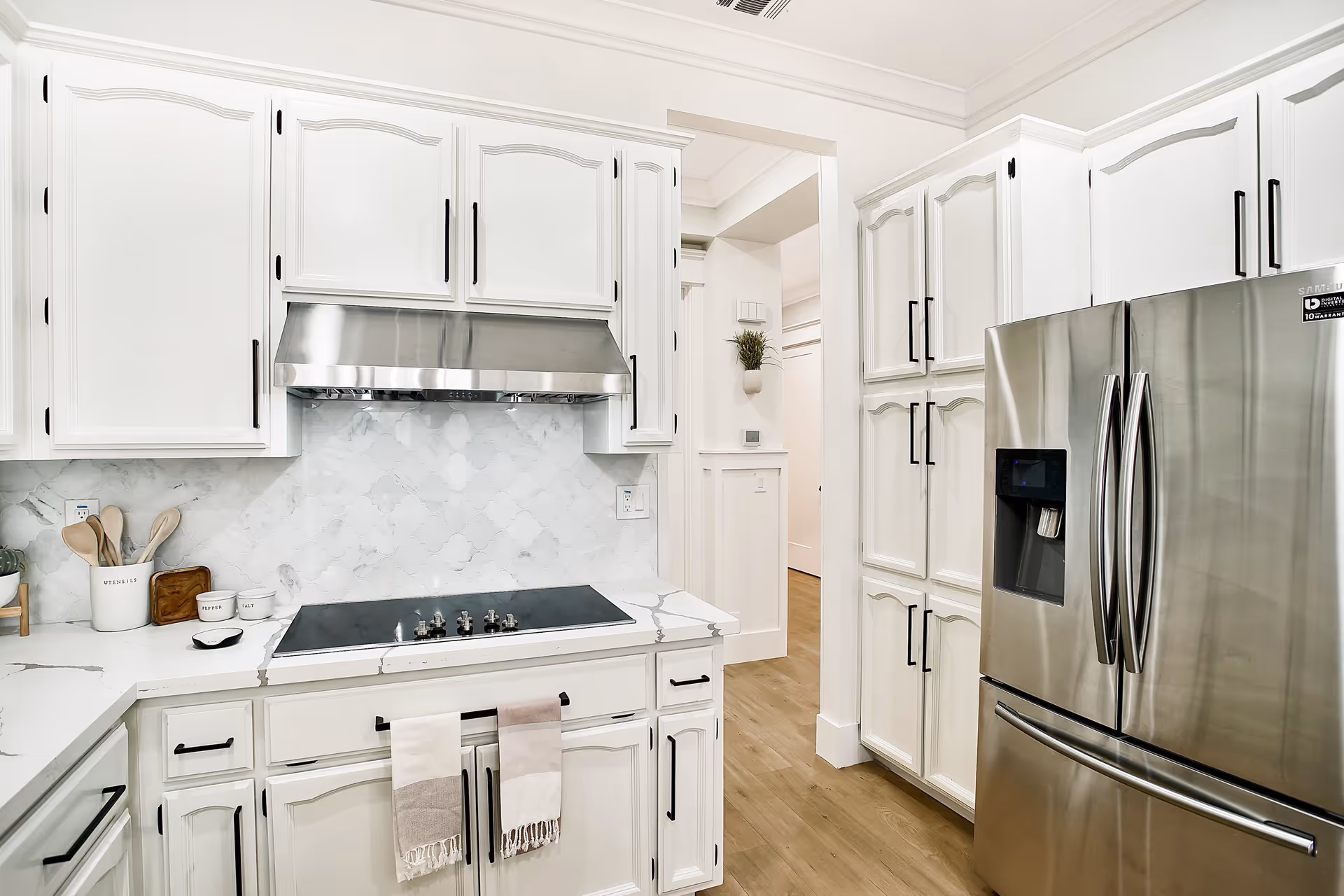 Bright and modern kitchen with white cabinetry, a stainless steel refrigerator, a stovetop with a stainless steel range hood, and a marble-patterned backsplash and countertop. Wooden utensils and small containers are placed on the counter, and two towels hang from the cabinet handles below the stovetop.