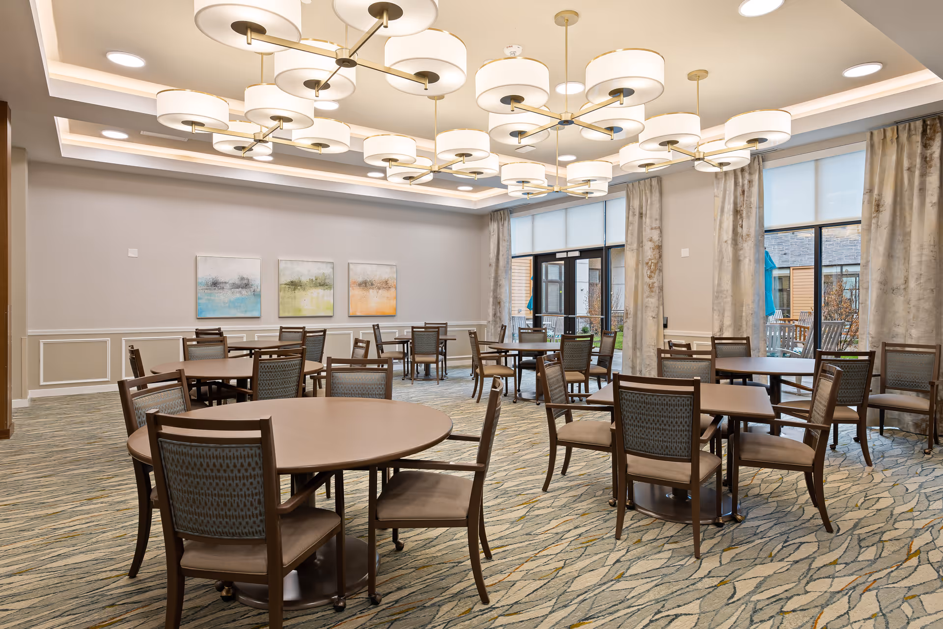 Spacious dining room with round and rectangular tables and chairs, large ceiling chandeliers, and tall windows leading outside.