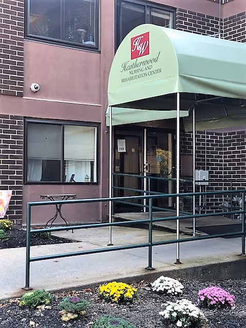 Entrance to Heatherwood Nursing and Rehabilitation Center with a green canopy over the doorway, a metal handrail along the ramp leading to the door, and a small garden with colorful flowers in front.