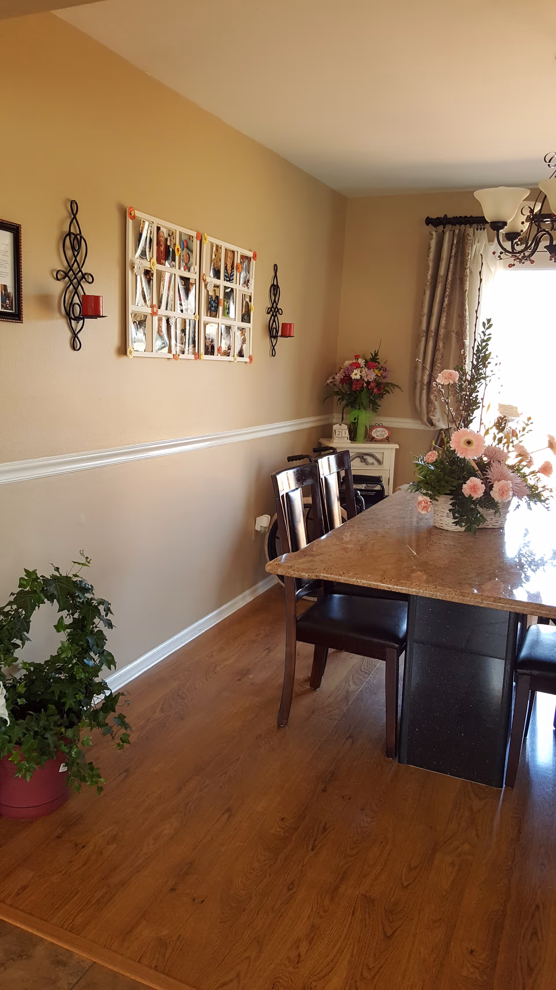 A dining room with a rectangular marble table surrounded by dark wooden chairs. There is a floral centerpiece on the table and a potted plant on the wooden floor. The walls are beige with white trim, decorated with framed photos and candle holders. A window with curtains allows natural light into the room.