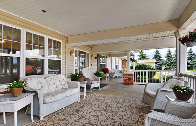 Covered front porch with wicker seating, potted plants, and a patterned rug overlooking landscaped grounds.
