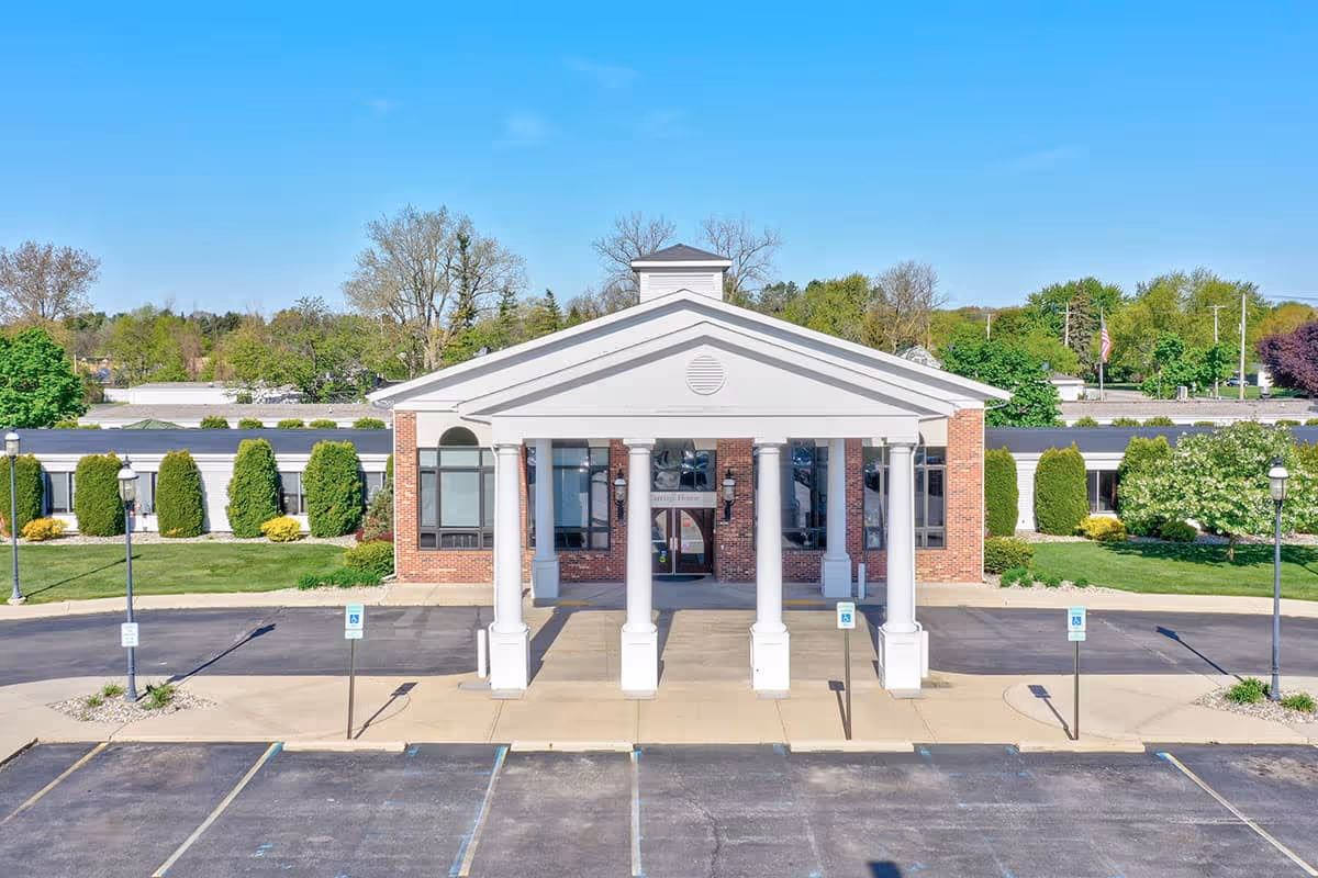 Front exterior view of Carriage House Nursing and Rehab building with a white portico supported by four columns, brick facade, large windows, and a paved parking area with handicap parking spaces in front. Green trees and shrubs surround the building under a clear blue sky.