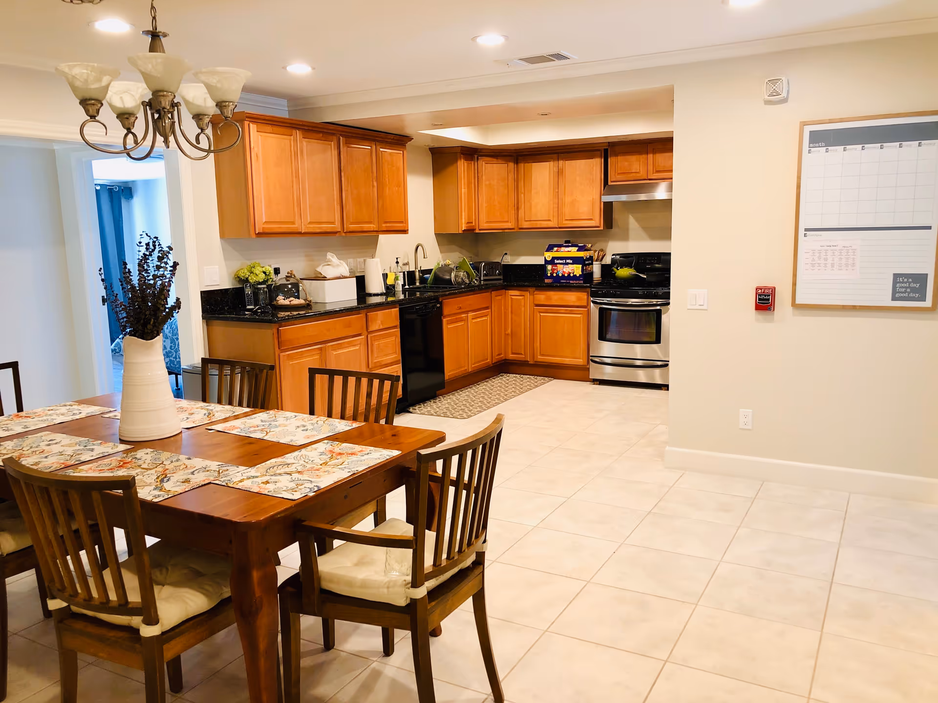A bright kitchen and dining area with wooden cabinets, black countertops, and stainless steel appliances. A wooden dining table with six chairs is set with floral placemats and a white vase with dried flowers. A chandelier hangs above the table, and a large whiteboard calendar is mounted on the wall.