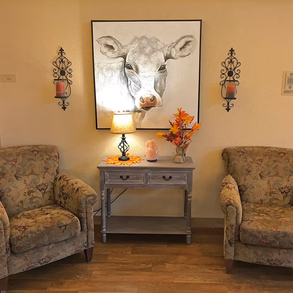 A cozy seating area with two floral upholstered armchairs on either side of a small gray wooden table. On the table, there is a lamp with a beige shade, a salt lamp, and a vase with autumn-colored leaves. Above the table hangs a large framed painting of a cow's face. Two decorative wall sconces with candles flank the painting on a beige wall. The floor is wooden.