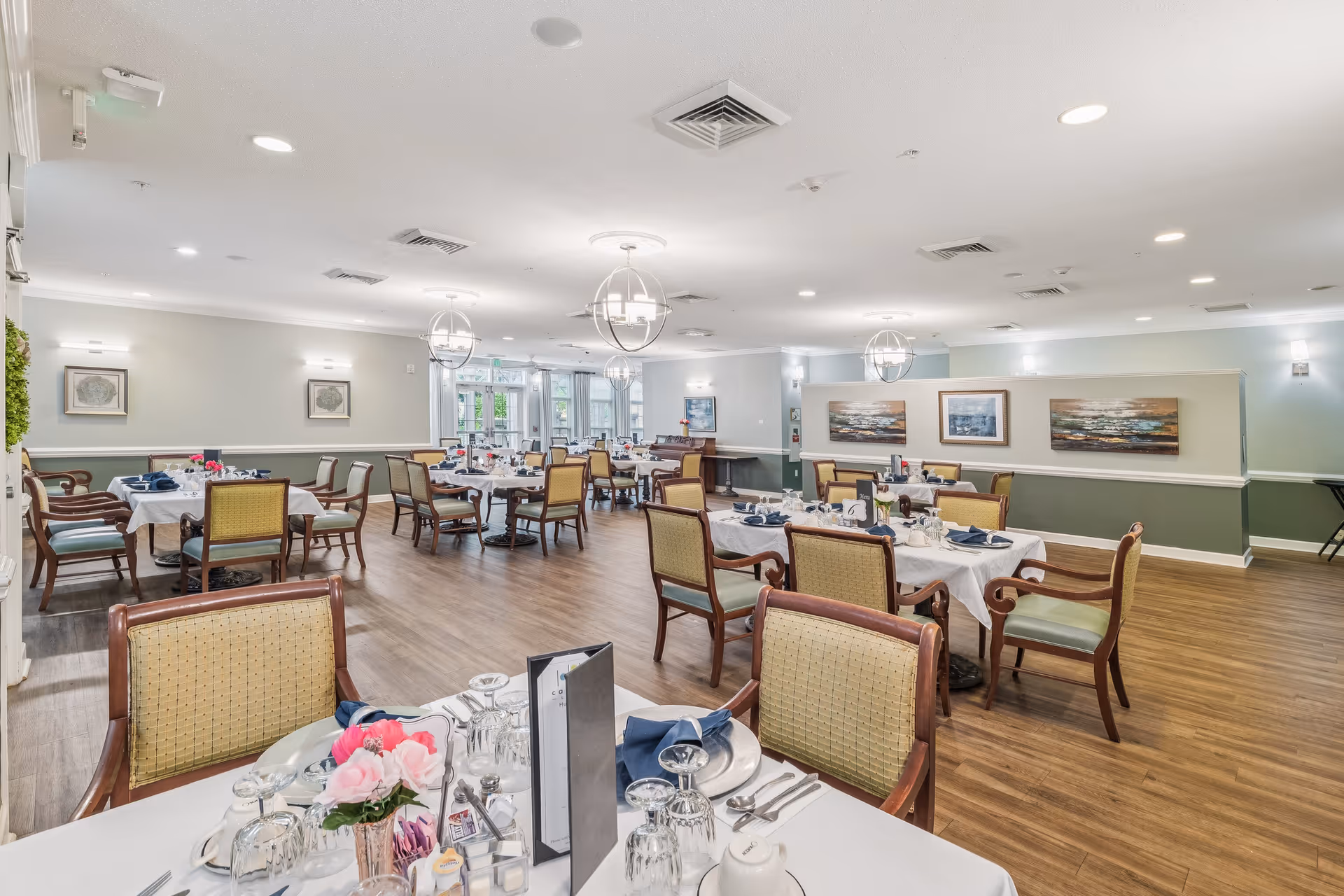 Bright, neatly set dining room with multiple tables and chairs and place settings in a senior living facility.