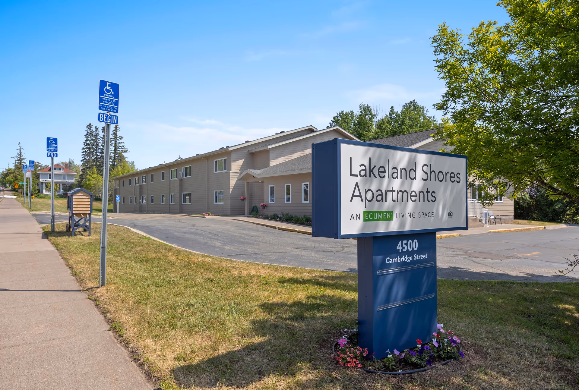 Exterior view of Lakeland Shores Apartments, a two-story beige building with multiple windows. In the foreground, there is a large sign displaying the name 'Lakeland Shores Apartments | An Ecumen Living Space' and the address '4500 Cambridge Street'. The area includes a sidewalk, grass, trees, and several handicap parking signs along the driveway.