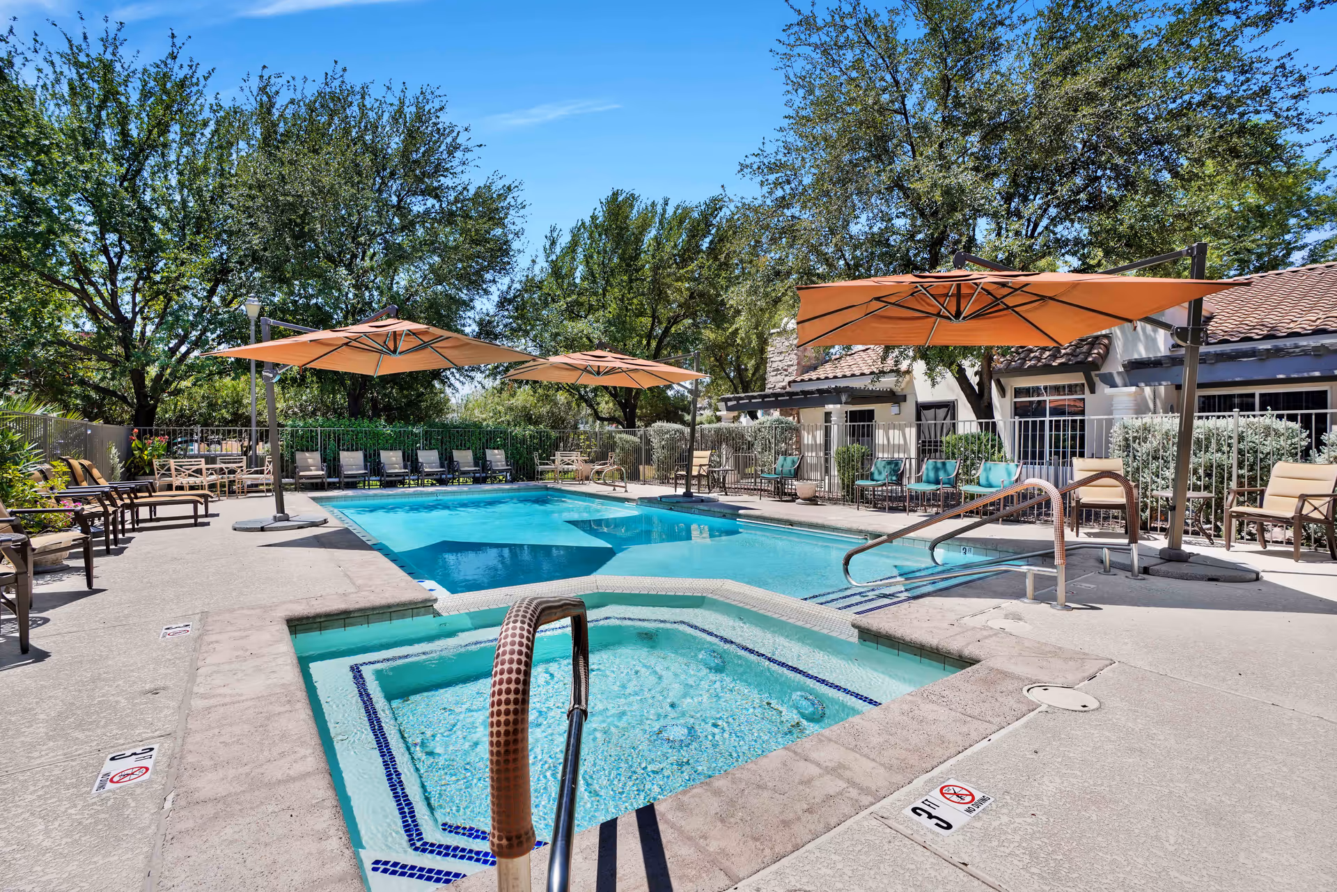 Outdoor swimming pool and adjacent hot tub with lounge chairs, large orange umbrellas, and a building in the background.