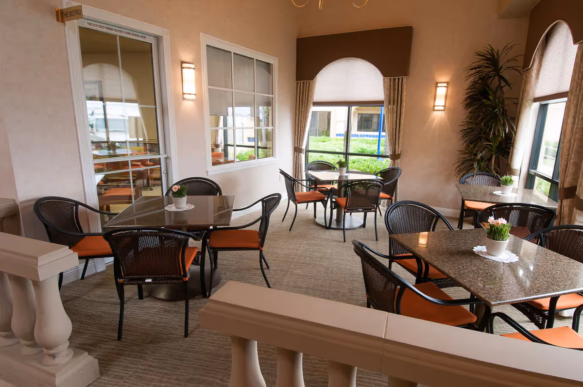 A cozy dining area with several square and round tables, each surrounded by black wicker chairs with orange cushions. Small potted plants are placed on each table. The room has large windows with beige curtains, allowing natural light to brighten the space. The walls are light-colored, and there are wall-mounted lights providing additional illumination. A door labeled 'The Bistro' is visible on the left side.