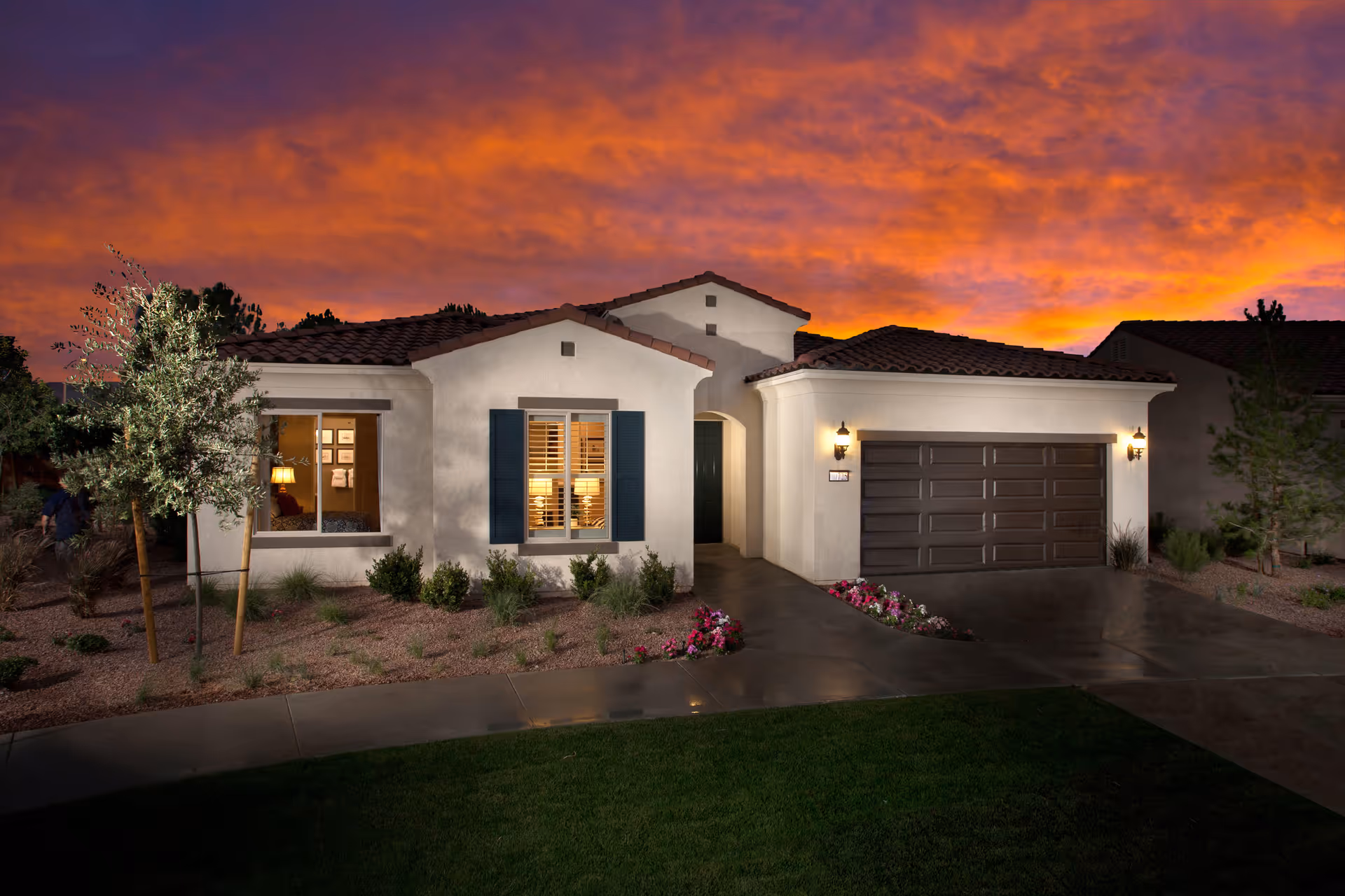 Exterior view of a single-story house at sunset with a dramatic orange and purple sky. The house features a tiled roof, a garage with a dark door, a front door under an archway, and windows with shutters. There are small bushes and flowers planted in the front yard with a concrete driveway and walkway.