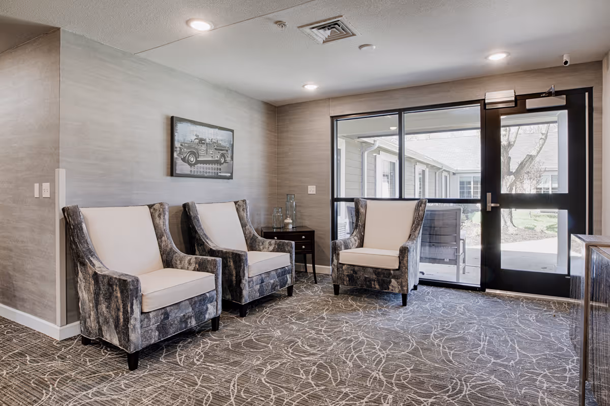 A small sitting area with three cushioned armchairs arranged around a dark wooden side table. The room has light gray walls, a patterned carpet, and a large glass door and window that provide a view of an outdoor courtyard with trees and a bench. A black and white framed picture of a vintage fire truck hangs on the wall.