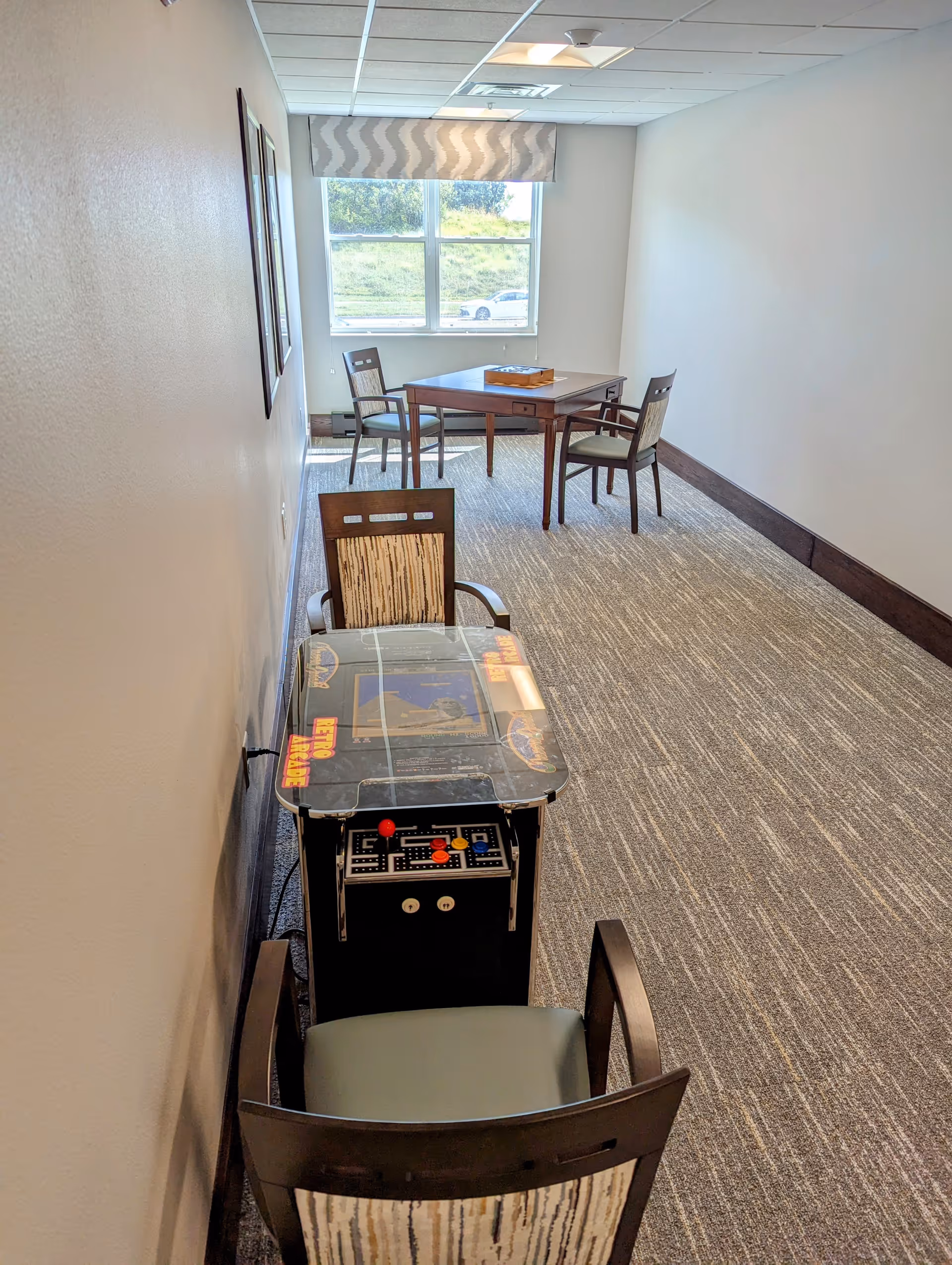 Long narrow interior activity room with a tabletop arcade game and several chairs leading to a table by a window.