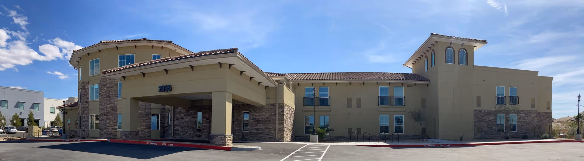 Exterior front view of a two-story senior living facility building with beige walls, stone accents, and a tiled roof under a blue sky with some clouds. The building has multiple windows and a covered entrance with the number 2895 displayed above it. There is a parking area and a fire lane in front of the building.