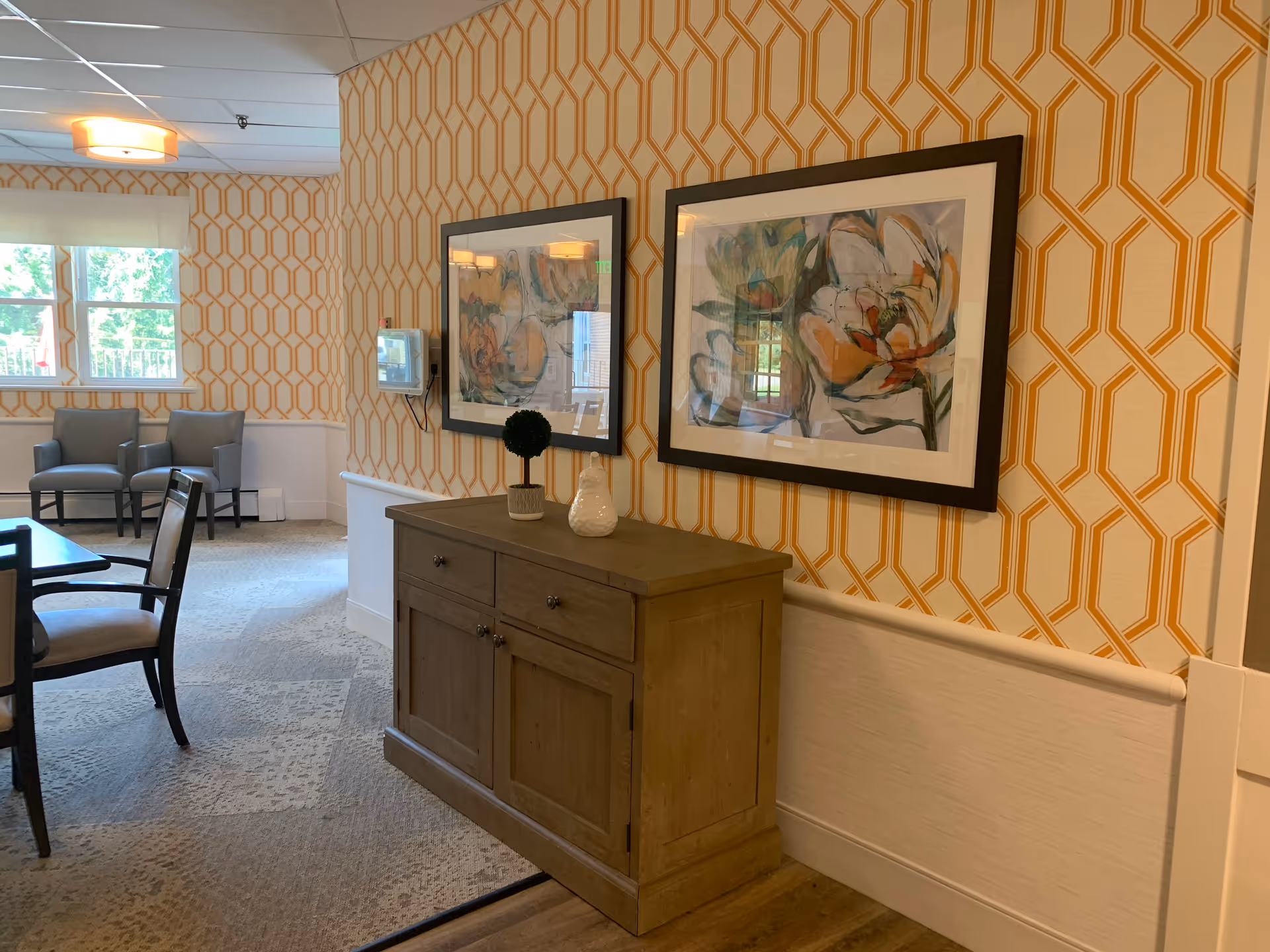Interior view of a common area in a senior living facility with patterned yellow and white wallpaper, a wooden cabinet with decorative items on top, two framed floral paintings on the wall, a table with chairs, and two armchairs near a window letting in natural light.