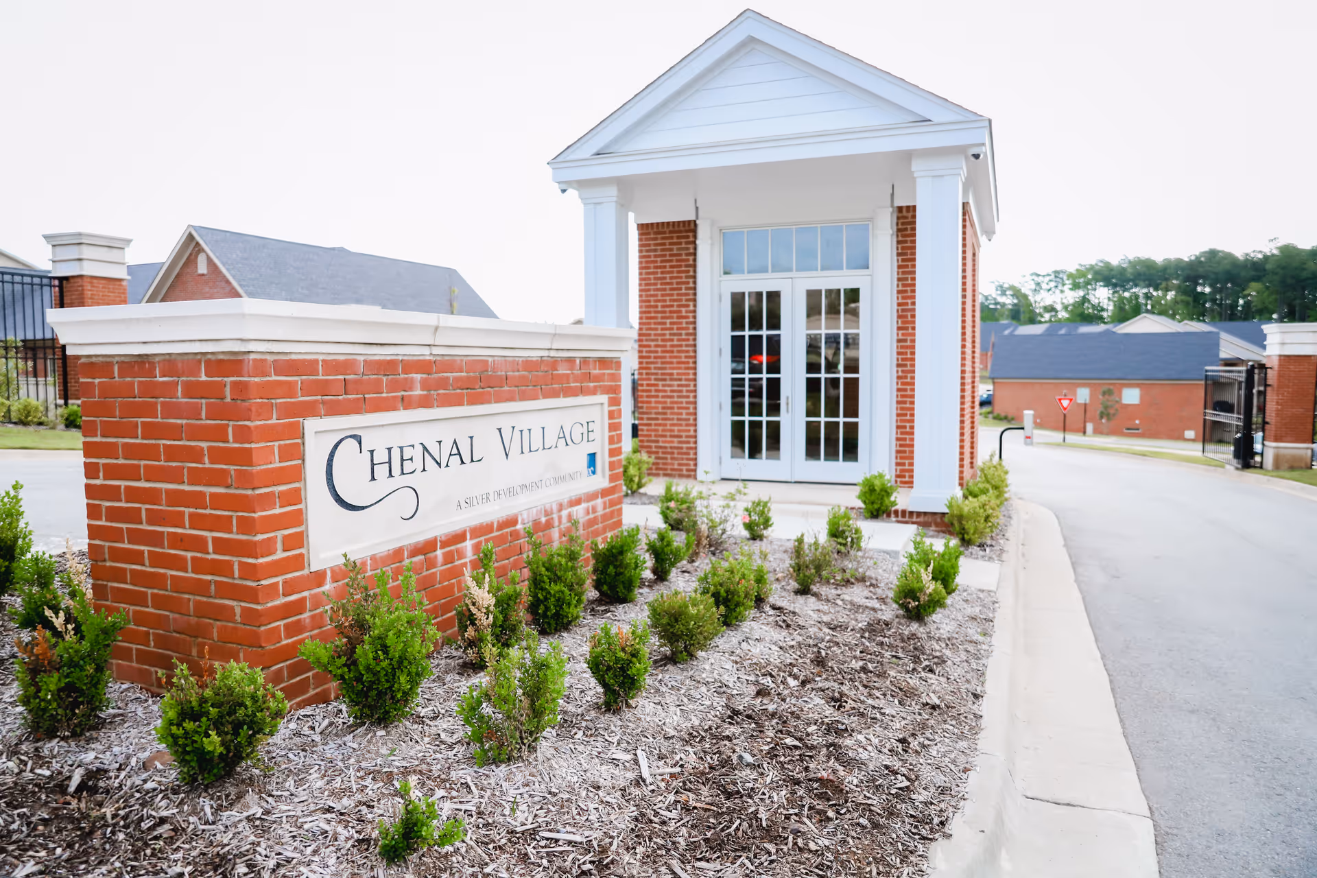 Entrance area of Chenal Village featuring a brick sign with the facility name and a small guardhouse or gatehouse with white columns and glass doors, surrounded by landscaping and a paved driveway.