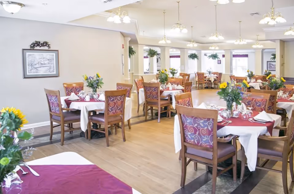 Bright dining room with multiple round tables set with red and white linens, wooden chairs, and floral centerpieces.