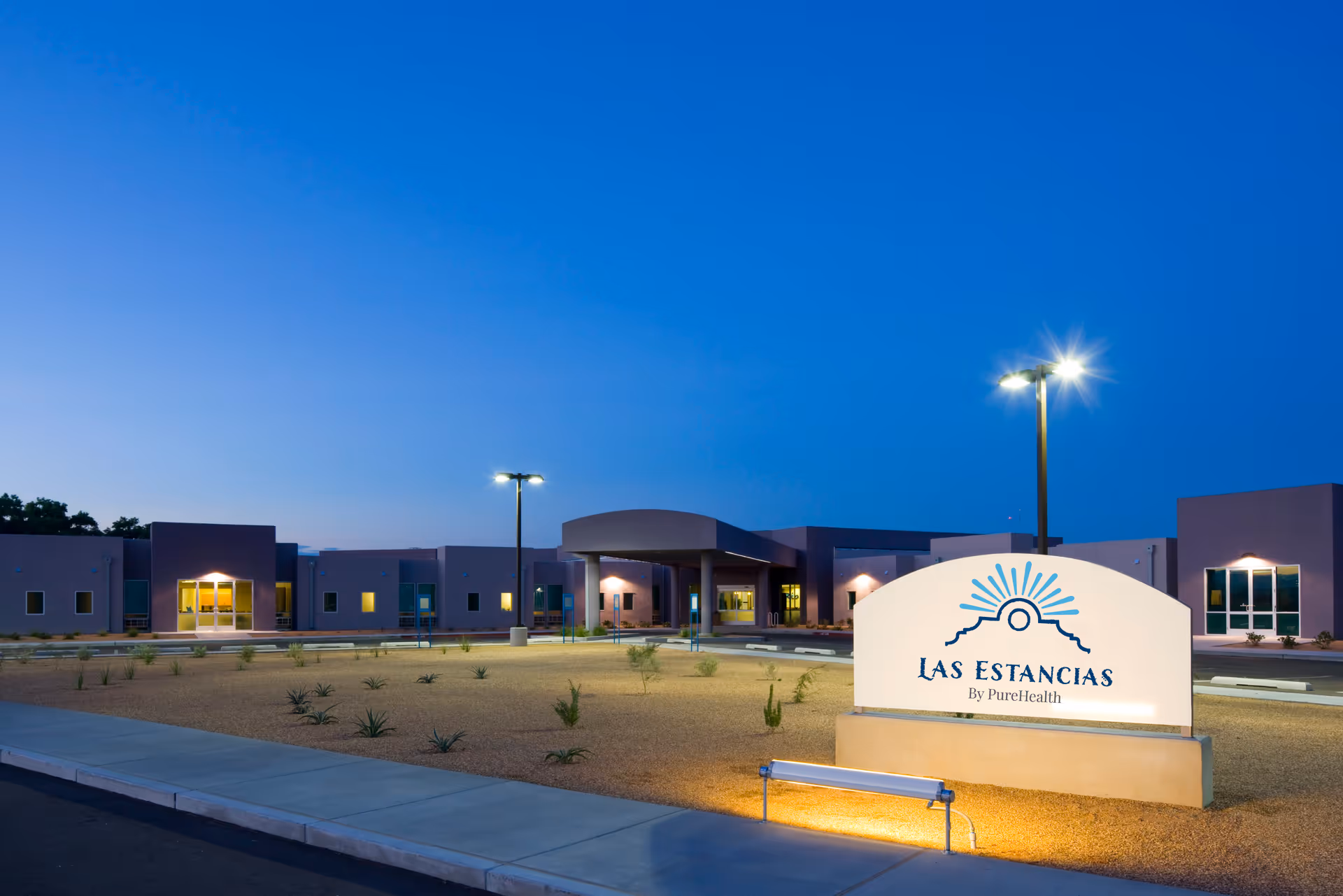 Exterior view of the Las Estancias facility at dusk with a clear blue sky, illuminated building entrance, and a lit sign displaying the facility name and logo in the foreground.