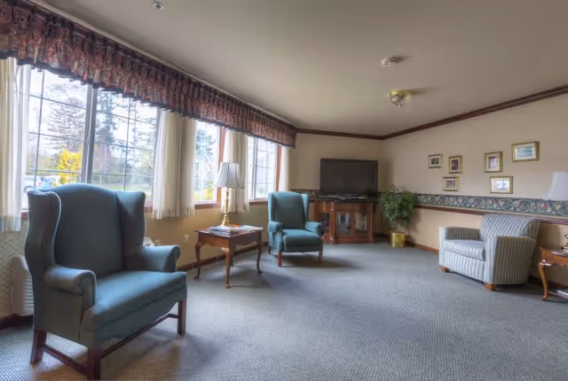 A bright sitting room with upholstered armchairs, side tables, and a television beneath large windows.