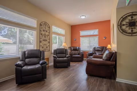 A cozy living room with five dark brown leather recliners arranged around the room. The walls are painted in warm beige and orange tones, with windows letting in natural light. There are two small lamps on side tables, a decorative wall clock, and a metal wall art piece.