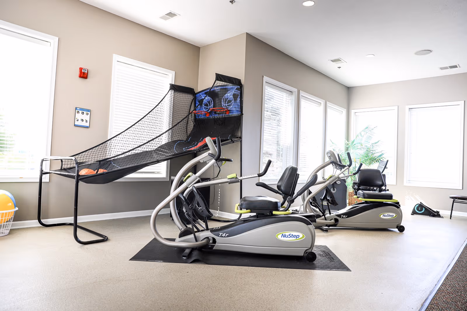 Bright exercise room with two NuStep recumbent cross trainers on black mats, a basketball arcade game, a basket with a large exercise ball, and several windows letting in natural light.