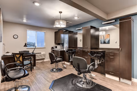 Interior view of a salon area in a senior living facility featuring two black salon chairs in front of large mirrors and dark wood cabinetry. There is a desk with a chair and a lamp near a window with blinds, and the room has wood flooring and neutral-colored walls with ceiling lights and a hanging light fixture.