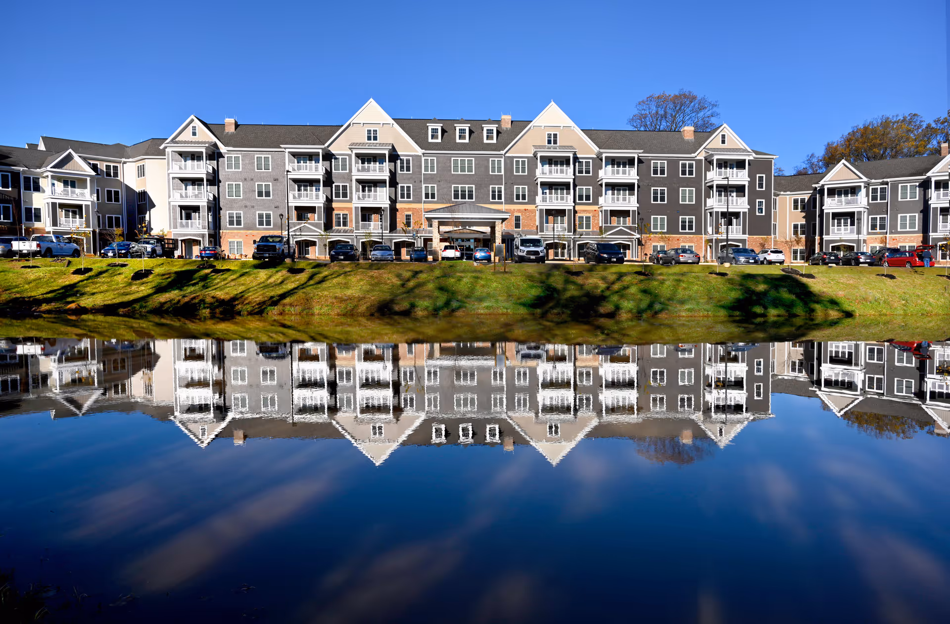 A large multi-story senior living community building with gray and beige exterior walls, multiple balconies, and a parking area in front. The building is reflected clearly in a calm body of water in the foreground under a bright blue sky.