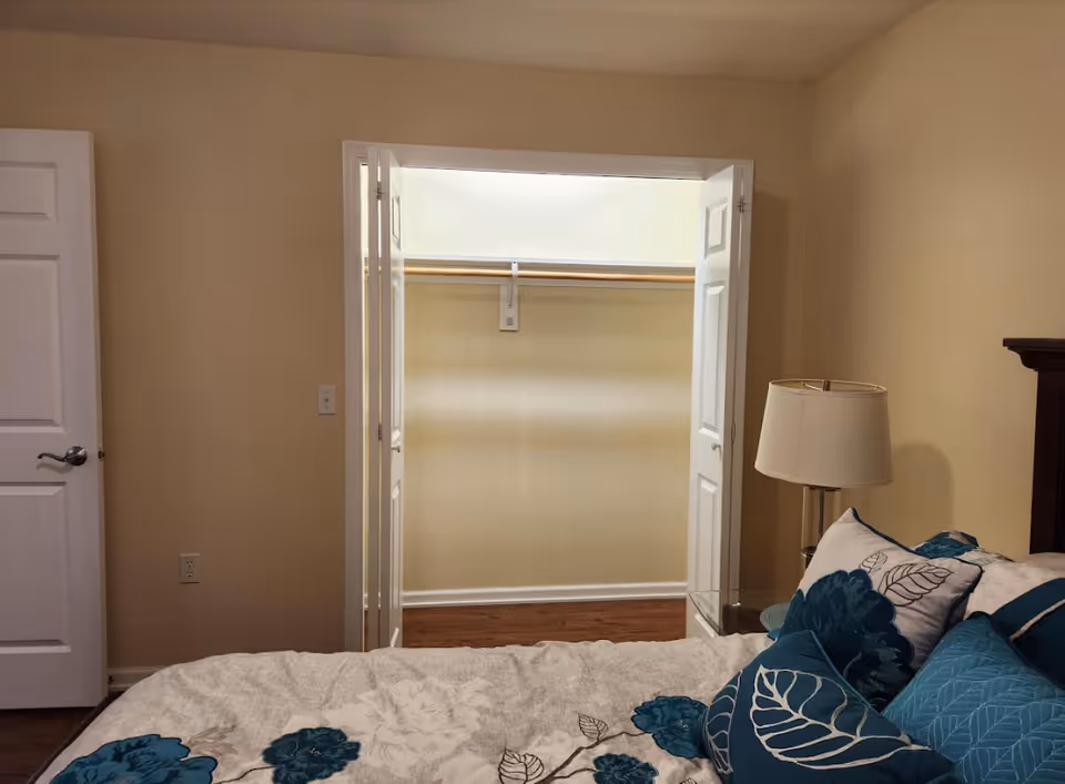 A bedroom with a bed covered in a floral patterned comforter and several pillows in shades of blue and white. To the right of the bed is a nightstand with a lamp. In the background, there is an open closet with double doors and a wooden floor.