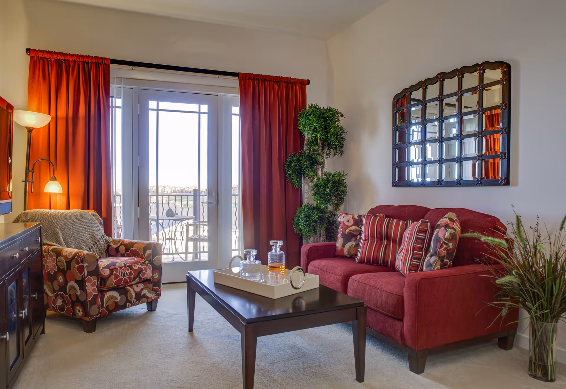 A cozy living room with a red sofa adorned with striped and floral cushions, a floral patterned armchair with a beige throw, a dark wooden coffee table with a tray holding glass decanters and a clock, a tall green plant in the corner, a large decorative mirror on the wall, and red curtains framing glass doors leading to a balcony with outdoor seating.