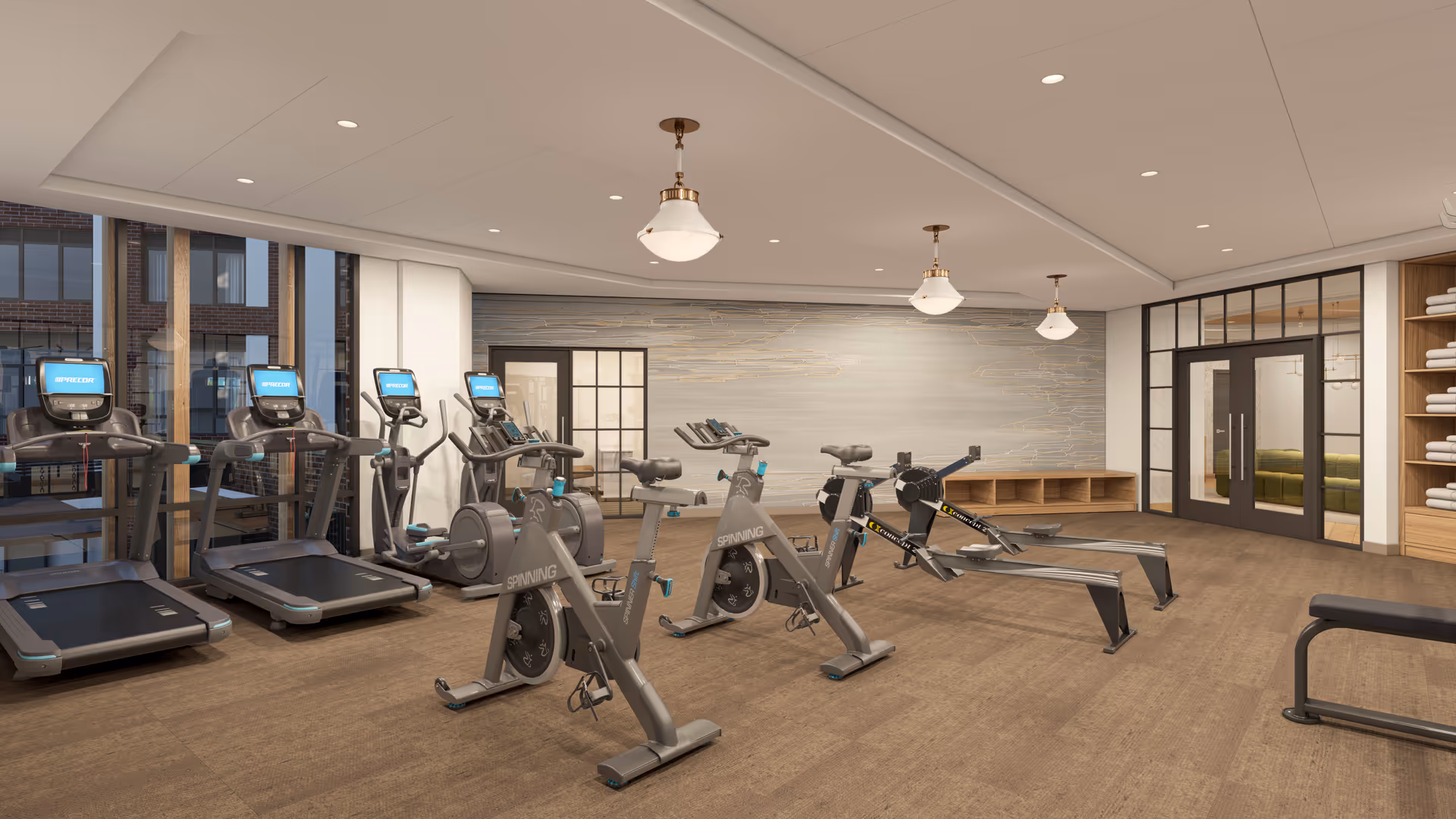 A modern fitness room with several exercise machines including treadmills, stationary bikes, and rowing machines. The room has large windows, wooden flooring, and a wall with a decorative design. Shelves with neatly folded towels are visible on the right side, and there are glass doors leading to another room.