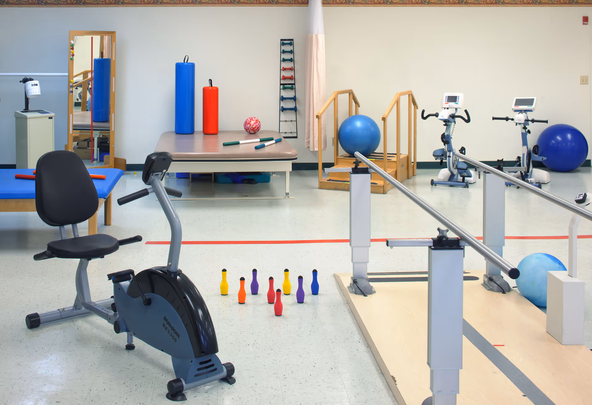 A physical therapy or rehabilitation room with exercise equipment including a stationary bike, parallel bars for walking practice, therapy balls, small colorful bowling pins, a padded table with foam rollers, and two exercise bikes in the background. There is also a mirror and wooden stairs for therapy exercises.