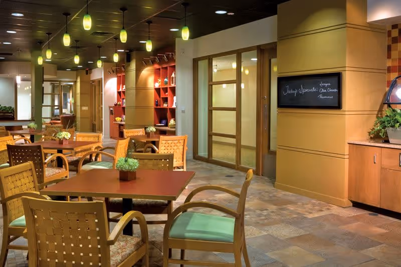 Interior view of a dining area in a senior living facility with wooden tables and chairs, green cushioned seats, pendant lights hanging from the ceiling, decorative plants on tables, and a wall with a framed menu board.