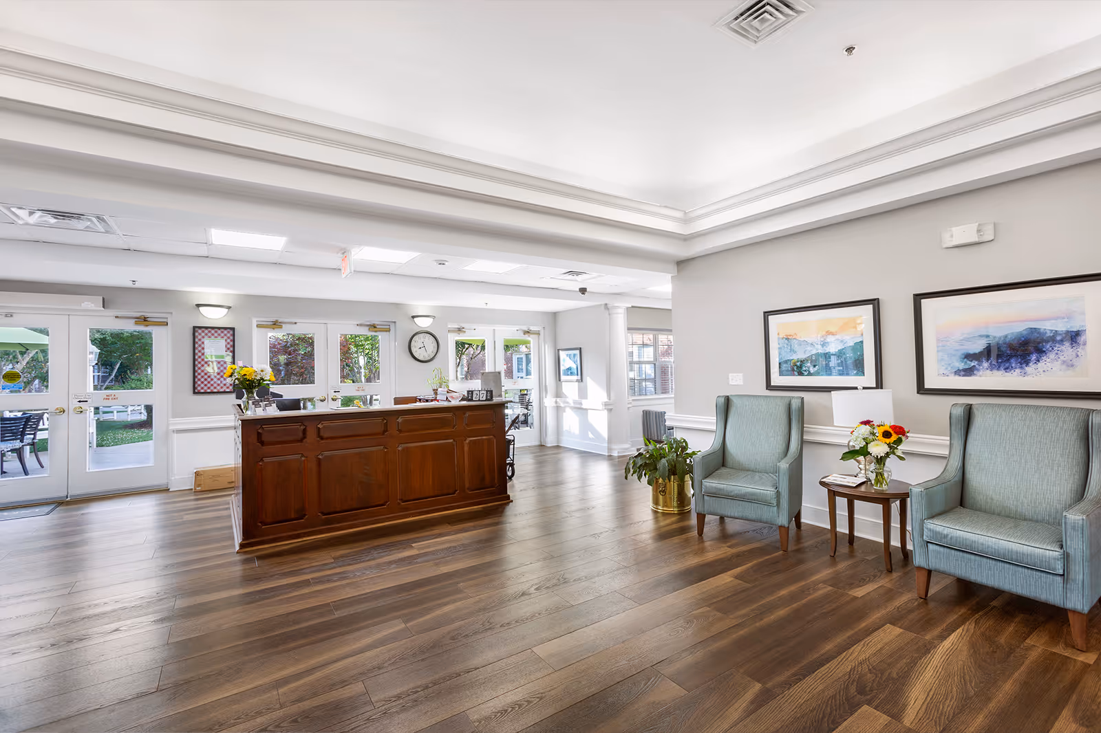 A bright and spacious reception area with a wooden front desk in the center. To the right, there are two light blue upholstered armchairs with a small wooden table between them holding a vase of flowers. The walls are light gray with two framed abstract paintings hanging above the chairs. The floor is a dark wood laminate, and there are multiple glass doors and windows letting in natural light, showing a glimpse of an outdoor patio area with green umbrellas and chairs.