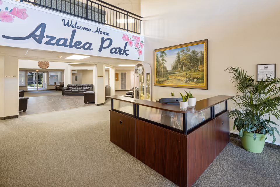 Reception area of Azalea Park senior living community with a wooden front desk, two small potted plants on the desk, a large framed landscape painting on the wall, a tall potted plant to the right, and a seating area with leather couches in the background. A large sign above reads 'Welcome Home Azalea Park' with pink flower decorations.