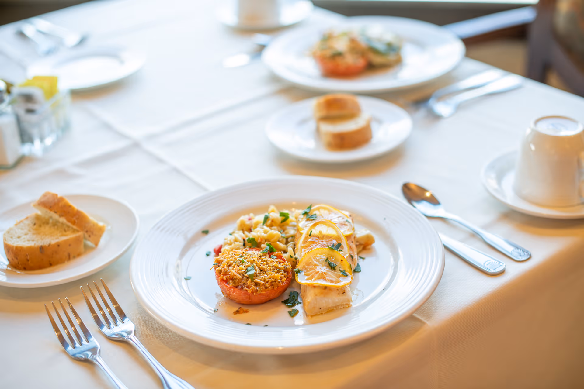 A plated meal of fish topped with lemon slices, a stuffed tomato and a side, served with bread, utensils and a cup on a white tablecloth in a dining setting.