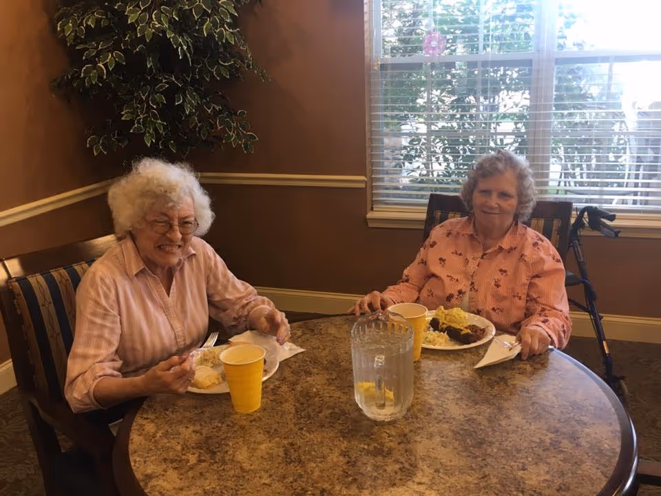 Two elderly women sitting at a round table in a dining area, eating a meal. One woman is wearing a light pink shirt and glasses, smiling, while the other woman is wearing a peach-colored shirt with a floral pattern and has a walker beside her. There is a pitcher of water and yellow cups on the table. A window with blinds and a green plant are visible in the background.