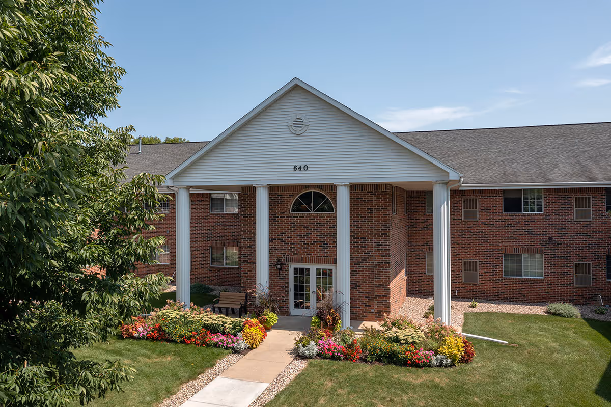 Front exterior view of a two-story brick apartment building with white columns supporting a triangular pediment above the entrance. The building number 640 is displayed on the pediment. There are colorful flower beds and green grass in front of the entrance, and a tree is visible on the left side under a clear blue sky.