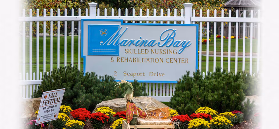 Outdoor view of a sign for Marina Bay Skilled Nursing & Rehabilitation Center located at 2 Seaport Drive, surrounded by a white fence, green bushes, and colorful flowers with a small sign advertising a Fall Festival event.