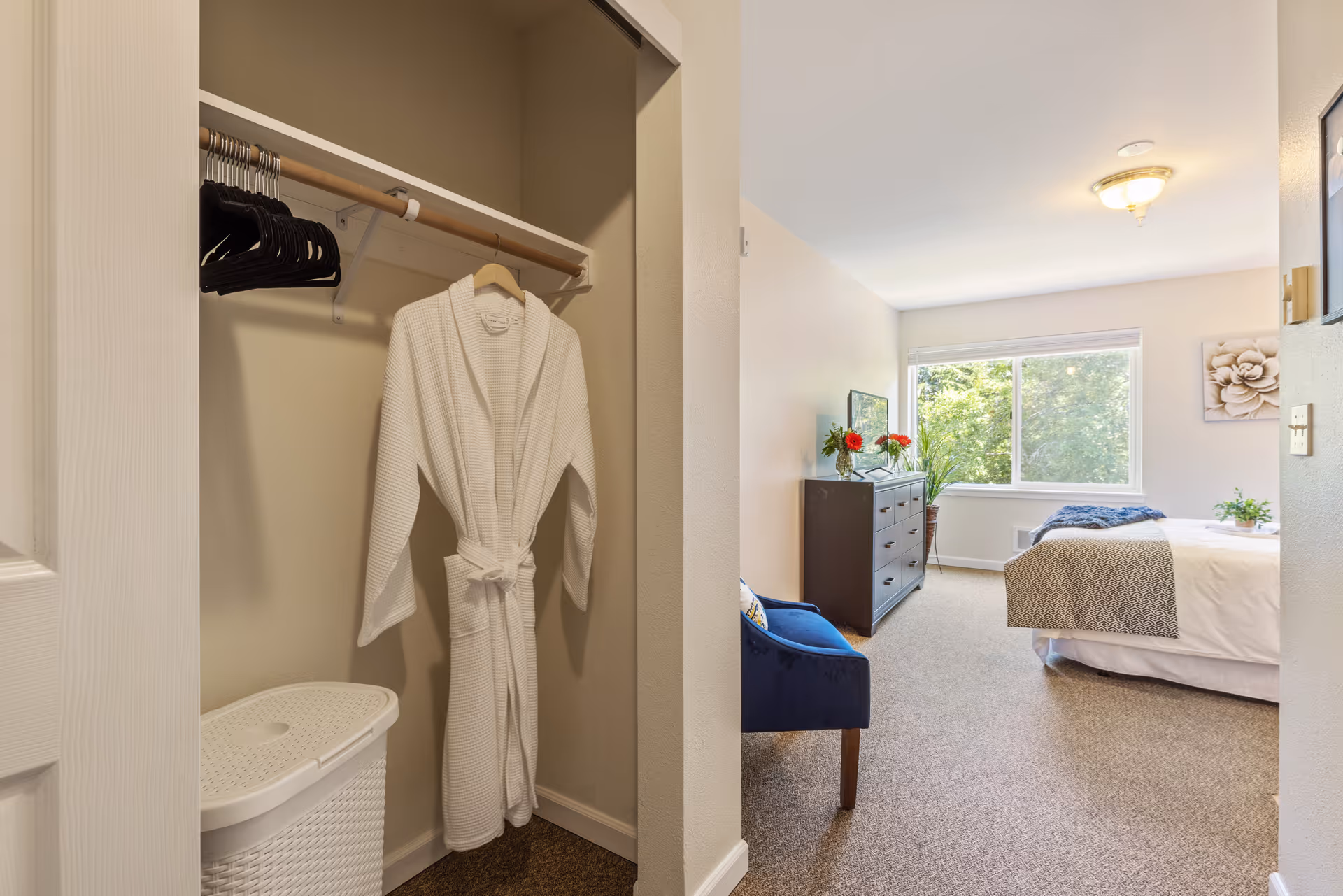 View of a bedroom from the entrance showing a closet with a white robe hanging and several black hangers. The bedroom has a bed with white and gray bedding, a dark wooden dresser with flowers on top, a blue upholstered chair, a large window with a view of greenery outside, and a wall decoration with a flower design.