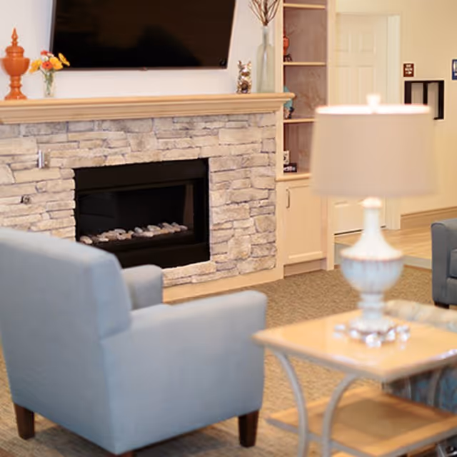 Comfortable lounge with a stone fireplace and wall-mounted TV, two blue armchairs, and a lamp-topped side table.