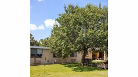 Single-story care facility building with a large tree and grassy lawn under a blue sky.