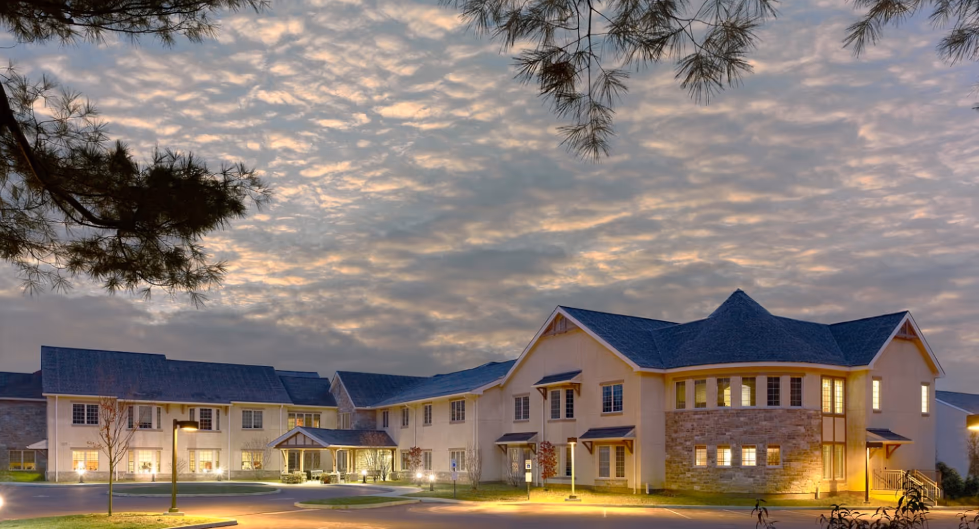 Exterior view of a two-story senior living facility building at dusk with lights on inside. The building has a combination of stone and light-colored siding with a peaked roof. There are trees and lamp posts in the parking area in front of the building, and the sky is partly cloudy with a soft evening glow.