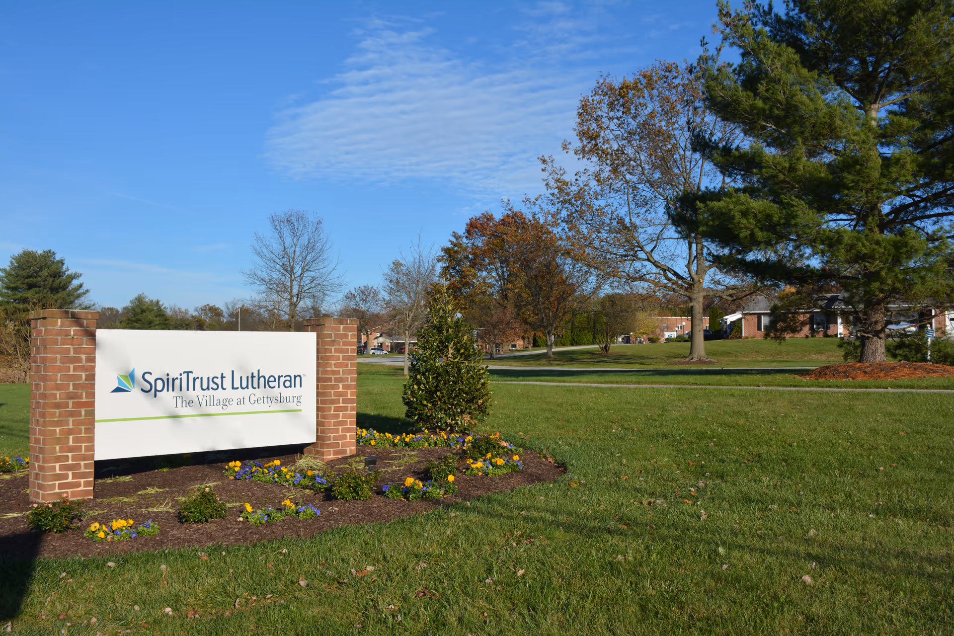 Outdoor view of SpiriTrust Lutheran, The Village at Gettysburg sign mounted on a brick structure surrounded by grass, flowers, and trees under a blue sky.